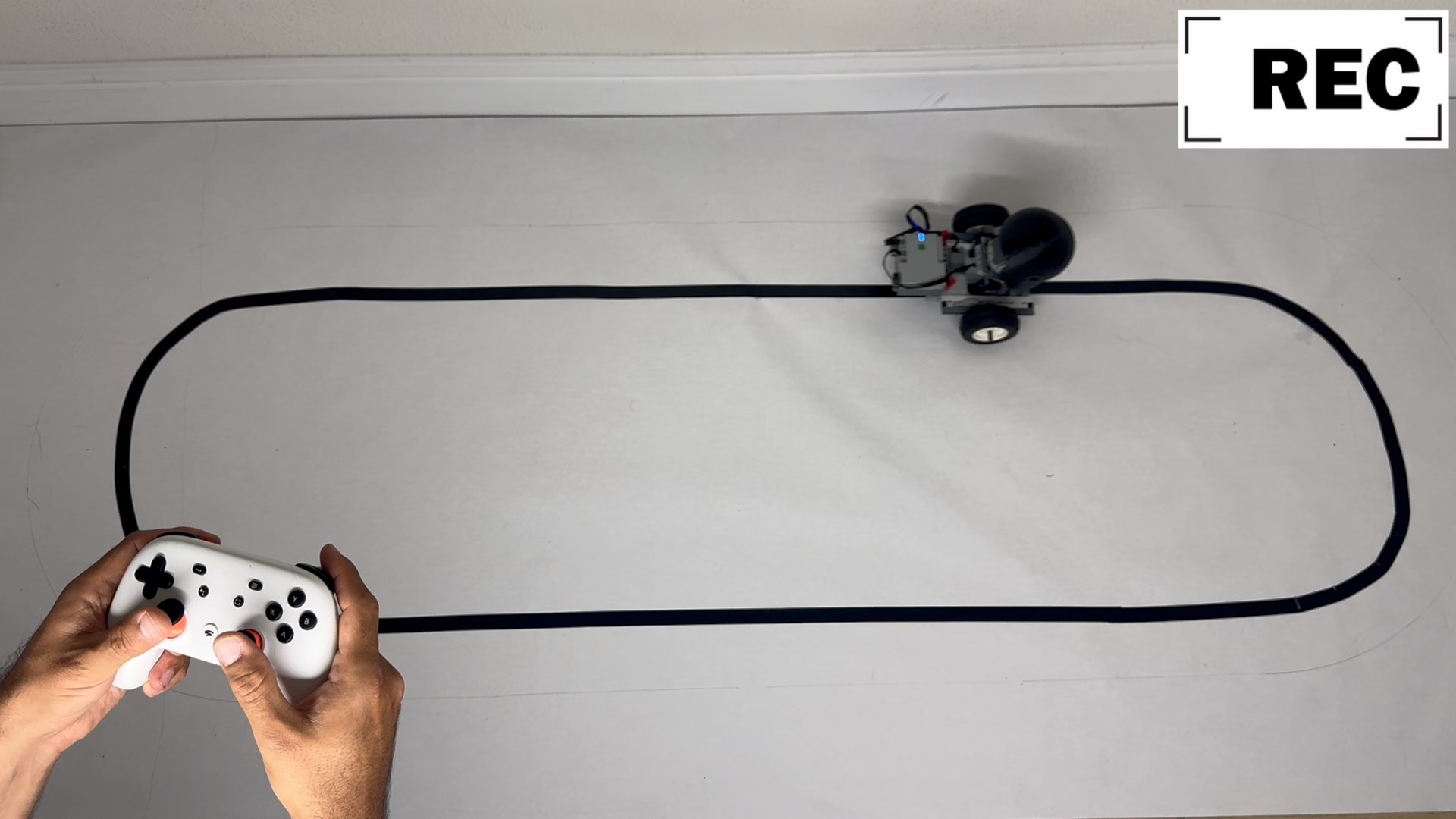 An operator drives Stack Chan around a black-tape loop with a Stadia controller while recording